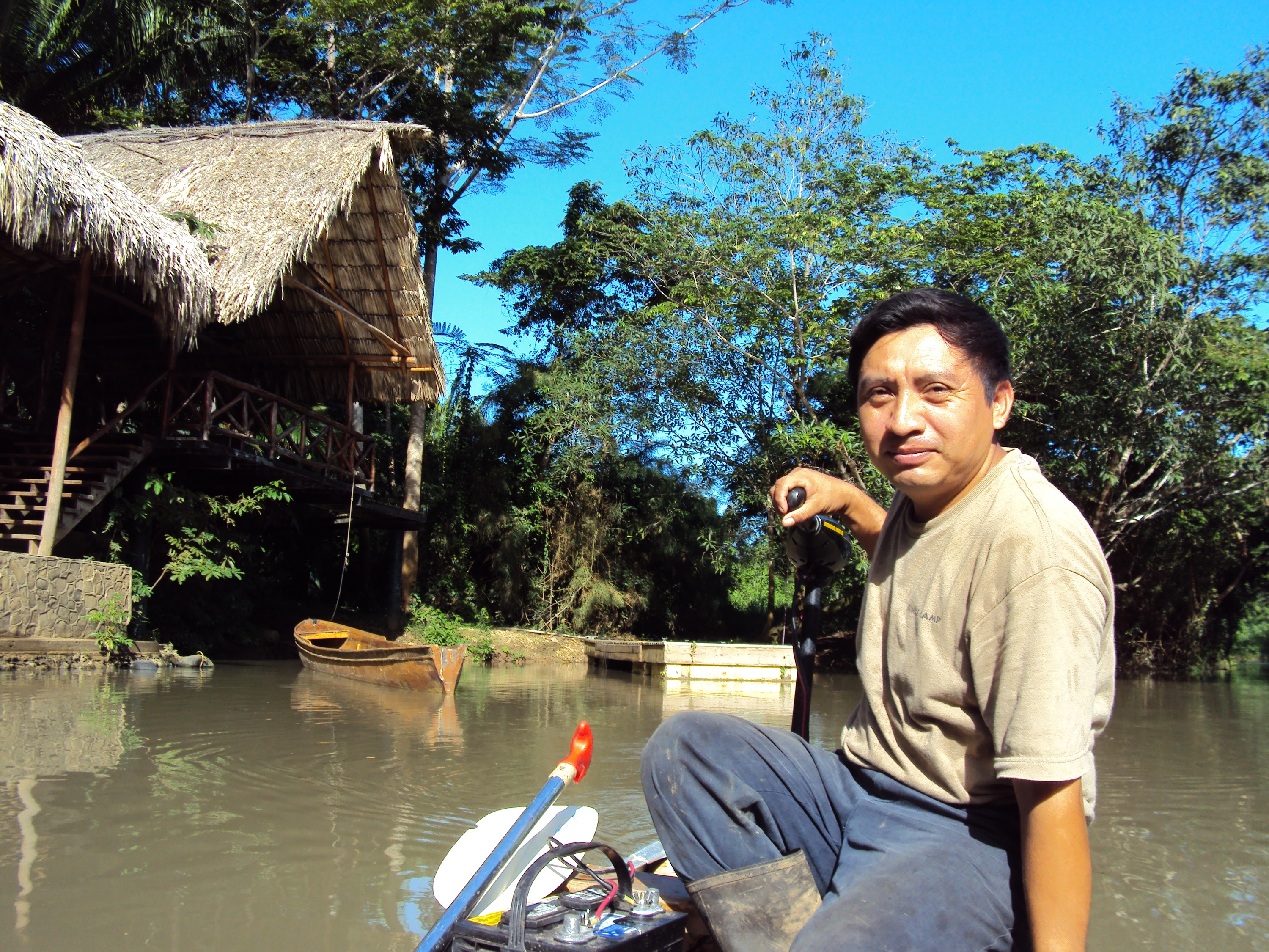 Man in a boat smiles surrounded by water and a thatched hut